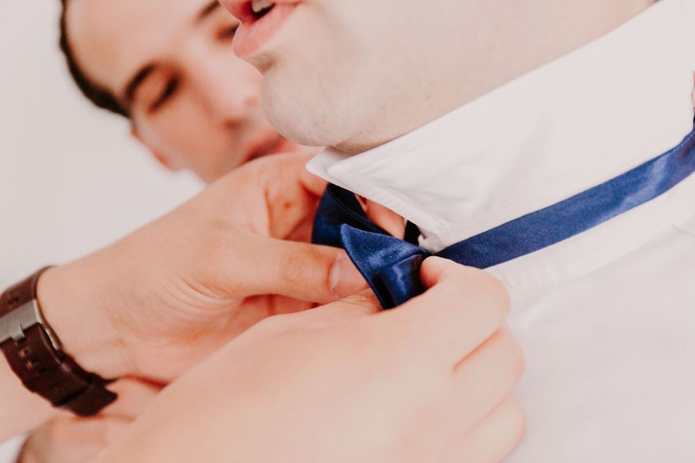 Close-up of hands helping groom tie blue bow tie on white dress shirt before wedding.