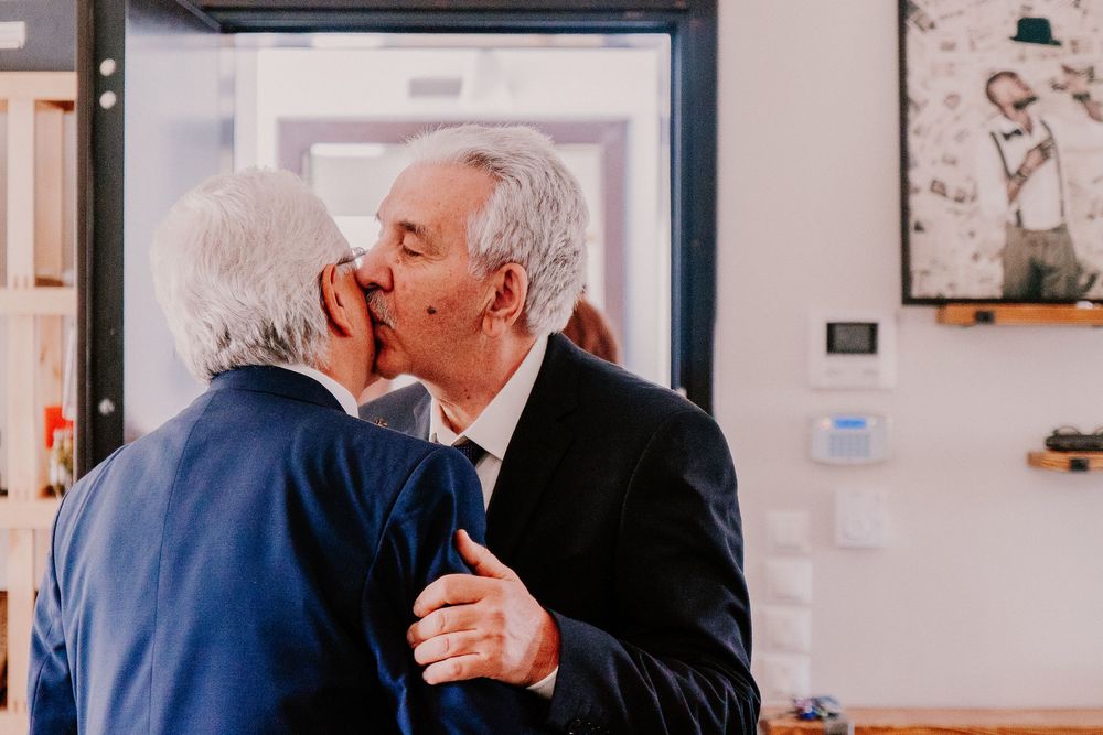 Two elderly men in suits sharing a kiss at a formal indoor event.
