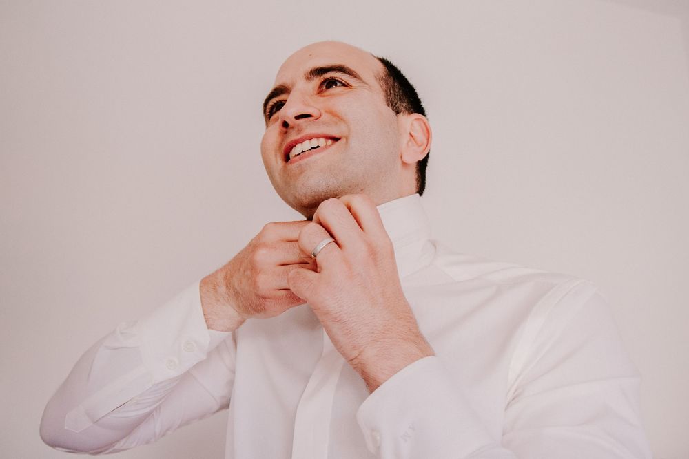 Smiling bald man in white dress shirt adjusting his collar button against a light background.