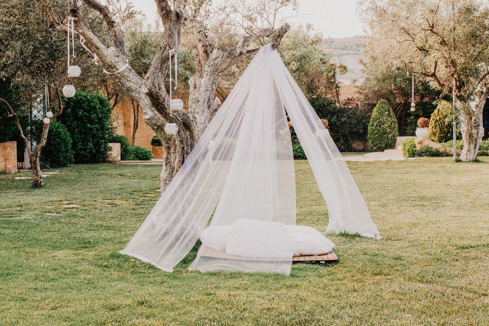 Elegant white sheer teepee tent with cushions set up in lush green garden for wedding.
