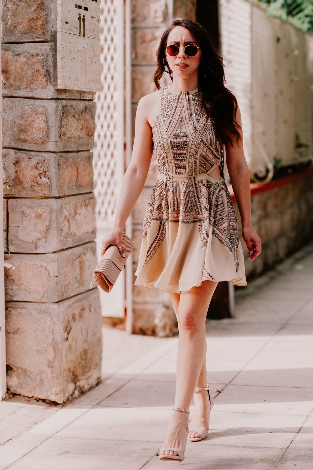 Woman in beige embroidered peplum dress and heels walking on a sunny street.