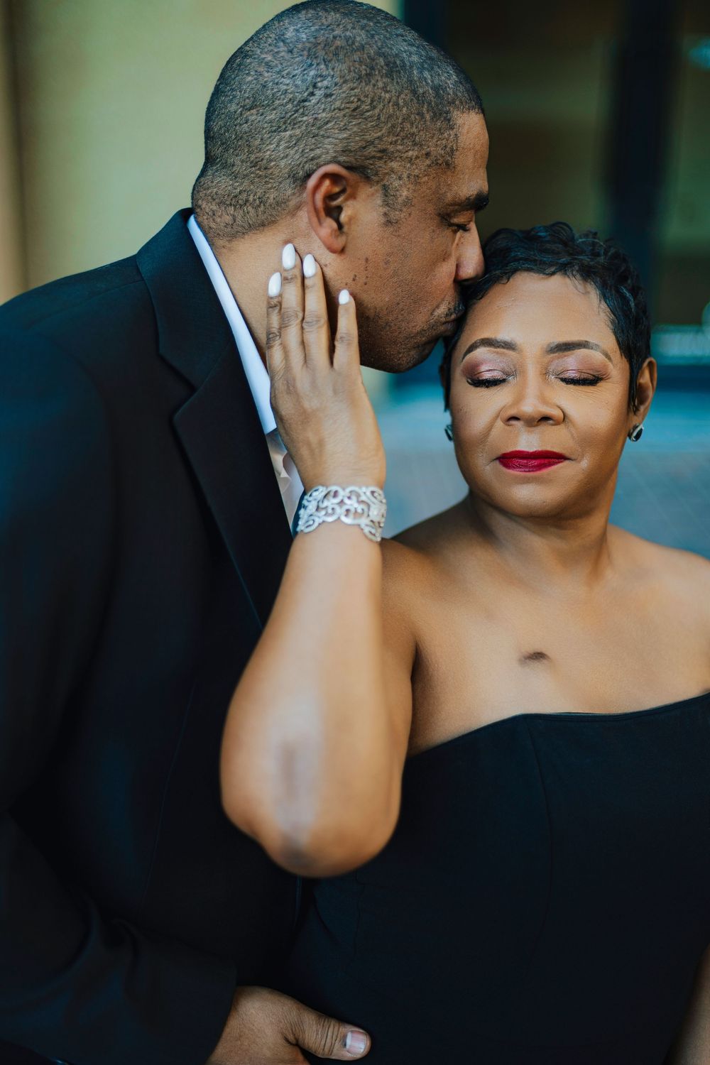 A man in a black suit tenderly kisses a woman's forehead as she closes her eyes, both dressed elegantly.