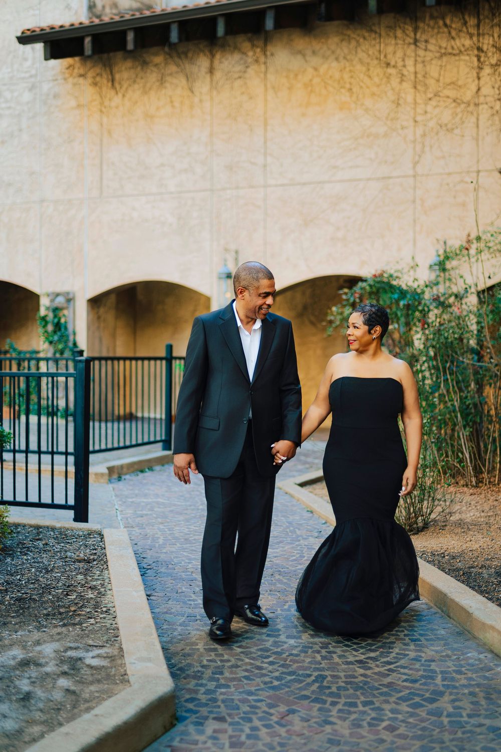 Elegant couple in black formal wear holding hands while walking along a cobblestone path near a building.