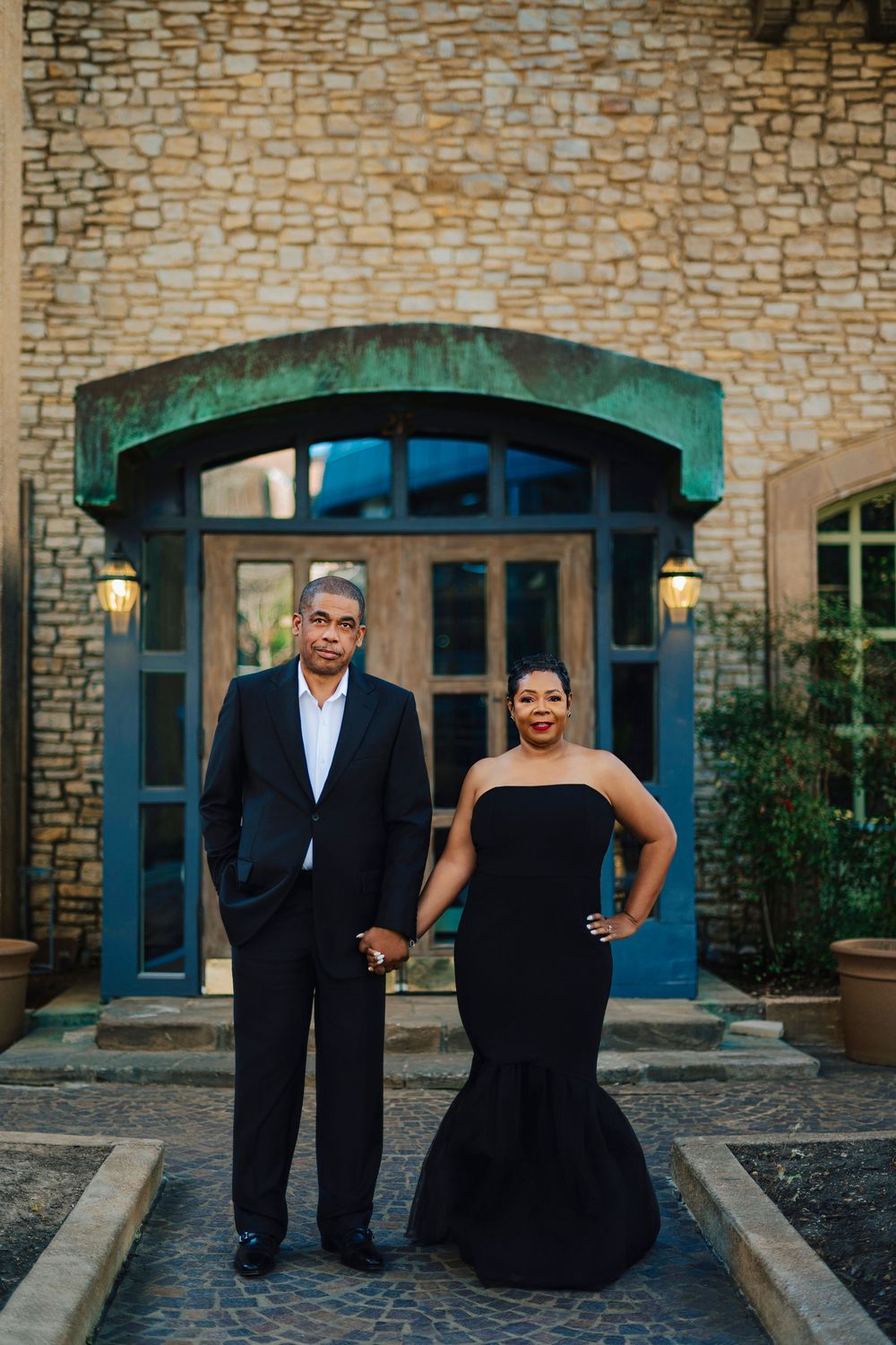 Elegant couple in matching black formal attire posing before a stone building with arched doorway.