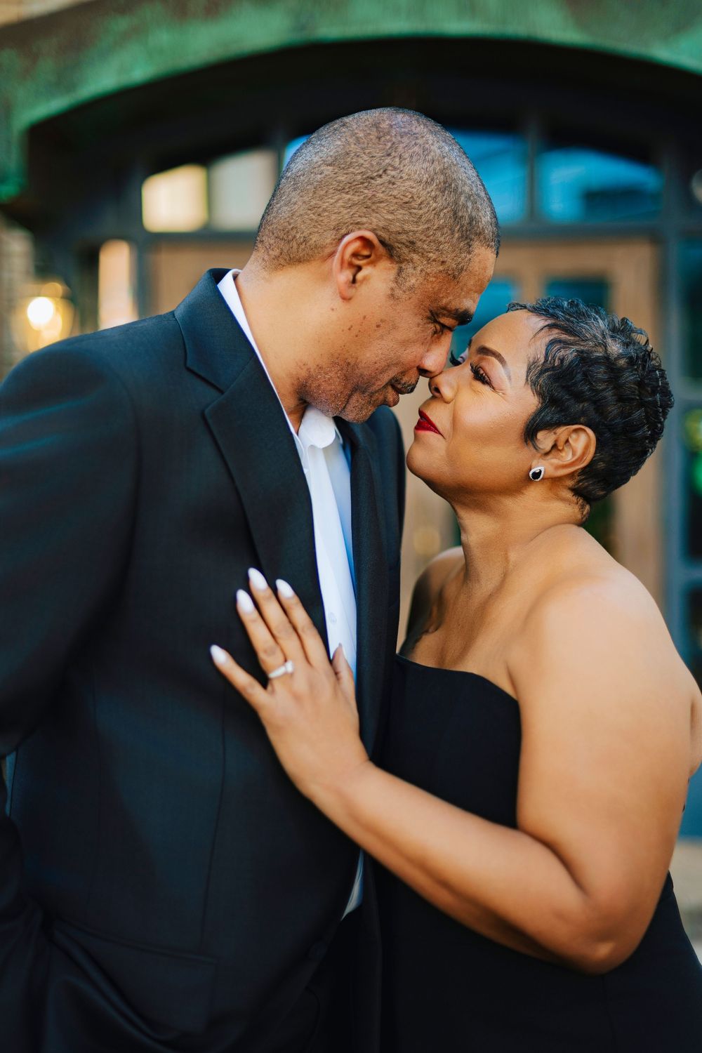 A couple in formal black attire share a romantic forehead touch, with the woman wearing an engagement ring.