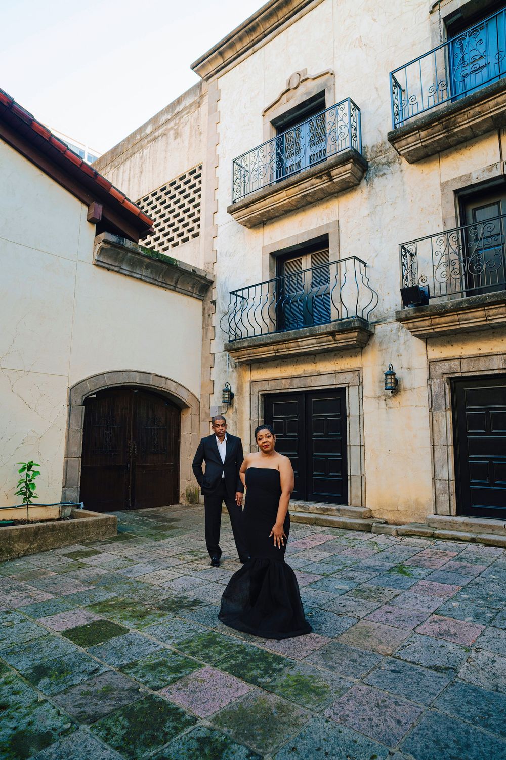 Elegant couple in black formal attire posing in a historic courtyard with ornate balconies and stone architecture.