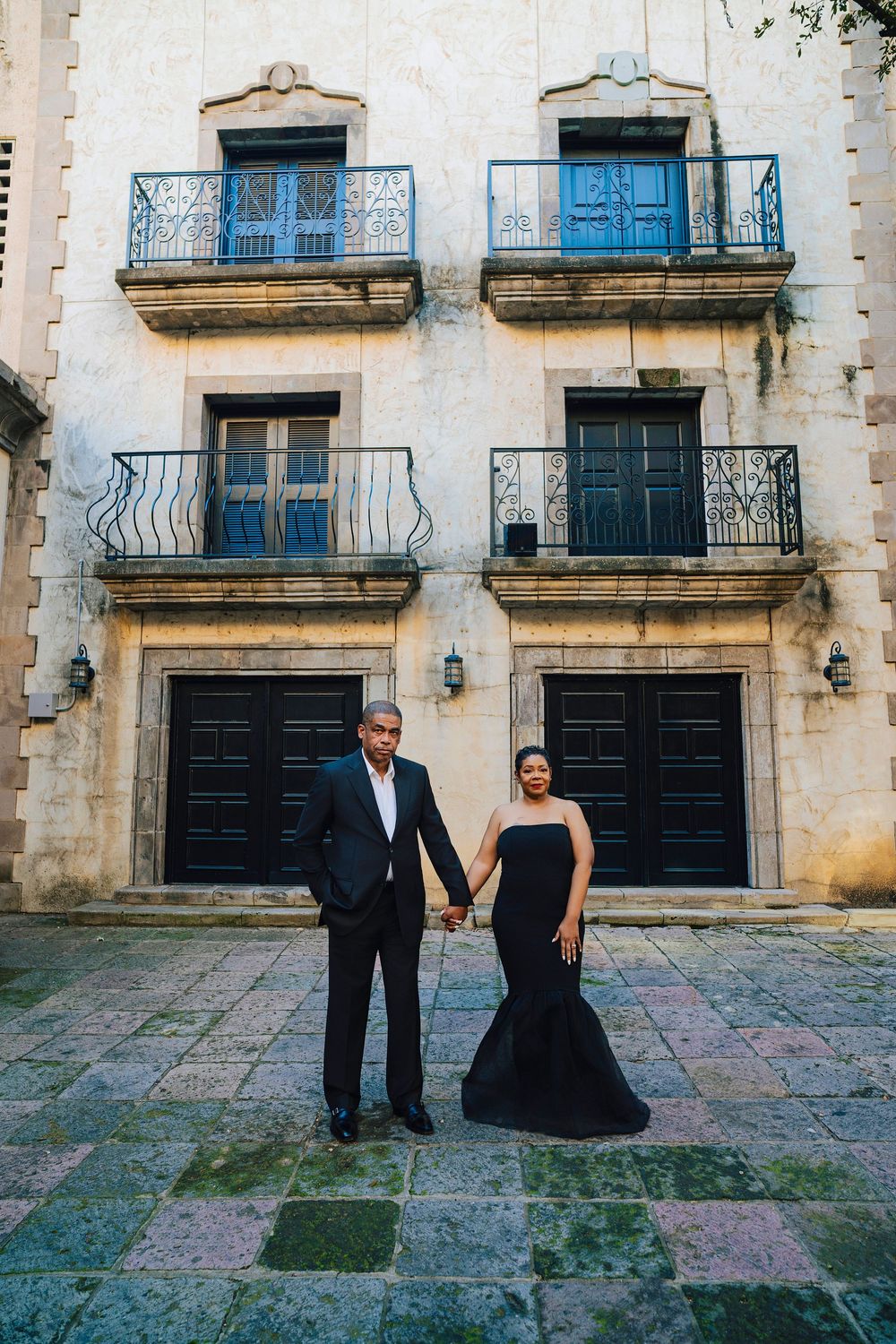 Elegant couple in black formal attire holding hands before a historic stone building with wrought iron balconies.