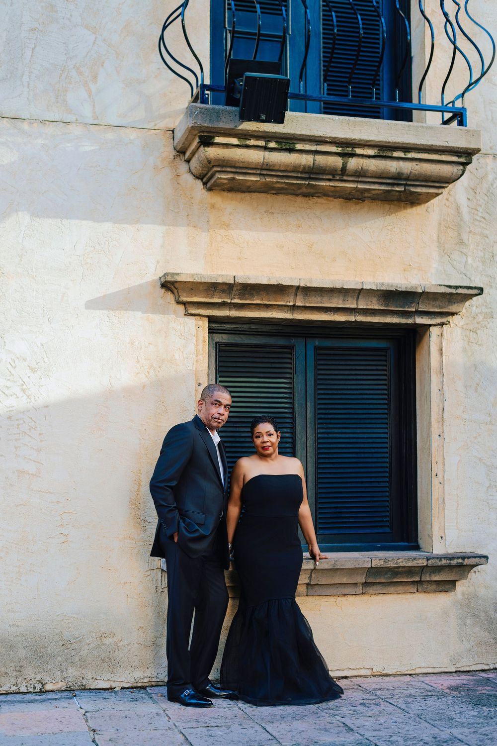 Elegant couple in matching black formal attire posing against a historic stone building with blue shuttered windows.