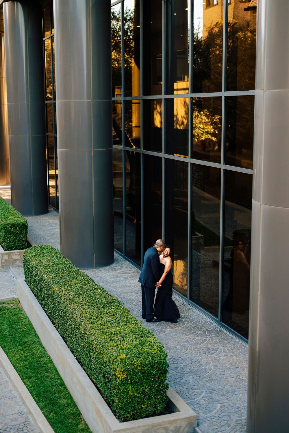 A couple shares a romantic embrace outside a modern glass building surrounded by manicured hedges at sunset.
