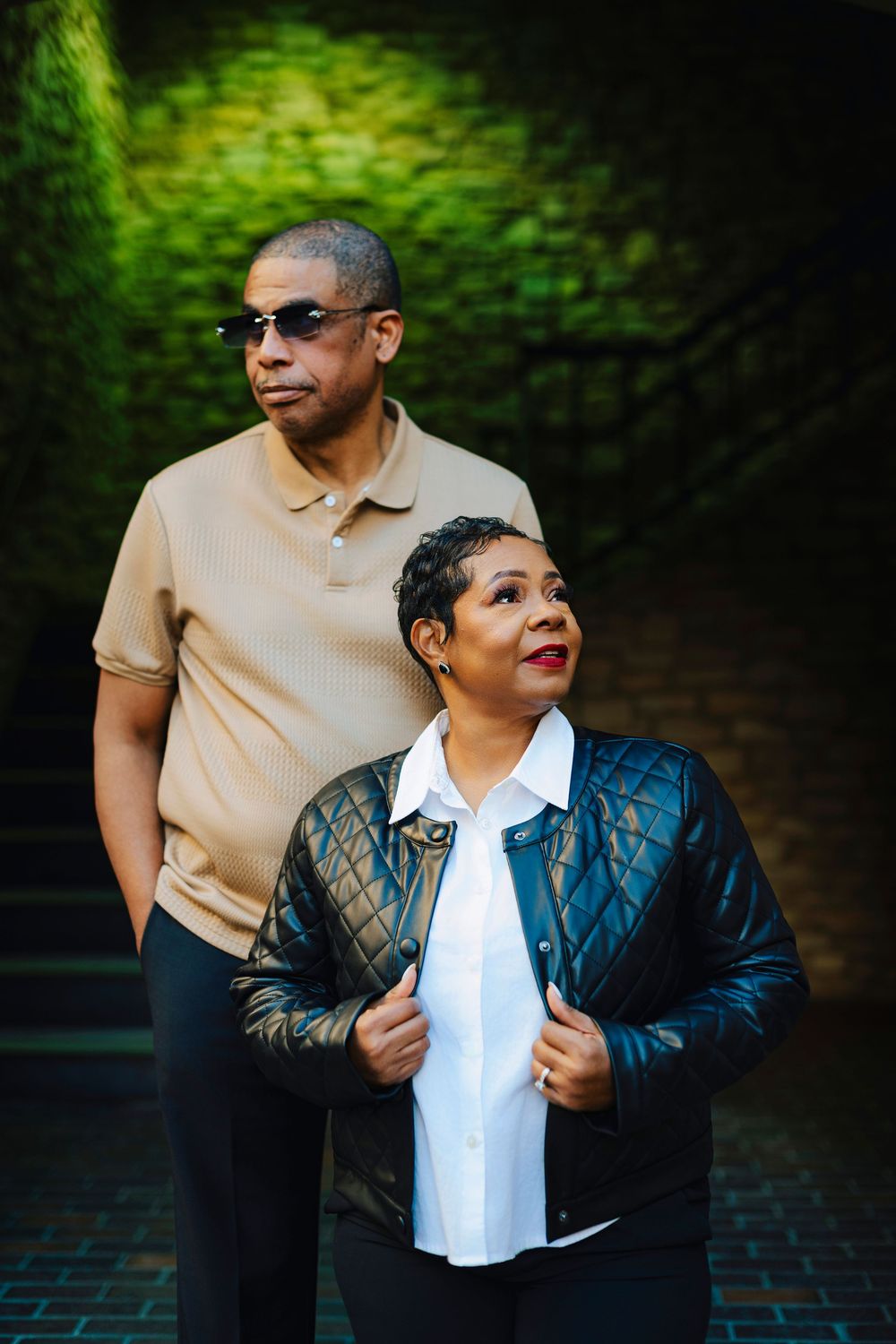 Stylish couple in casual chic outfits posing against a lush green ivy-covered wall backdrop.