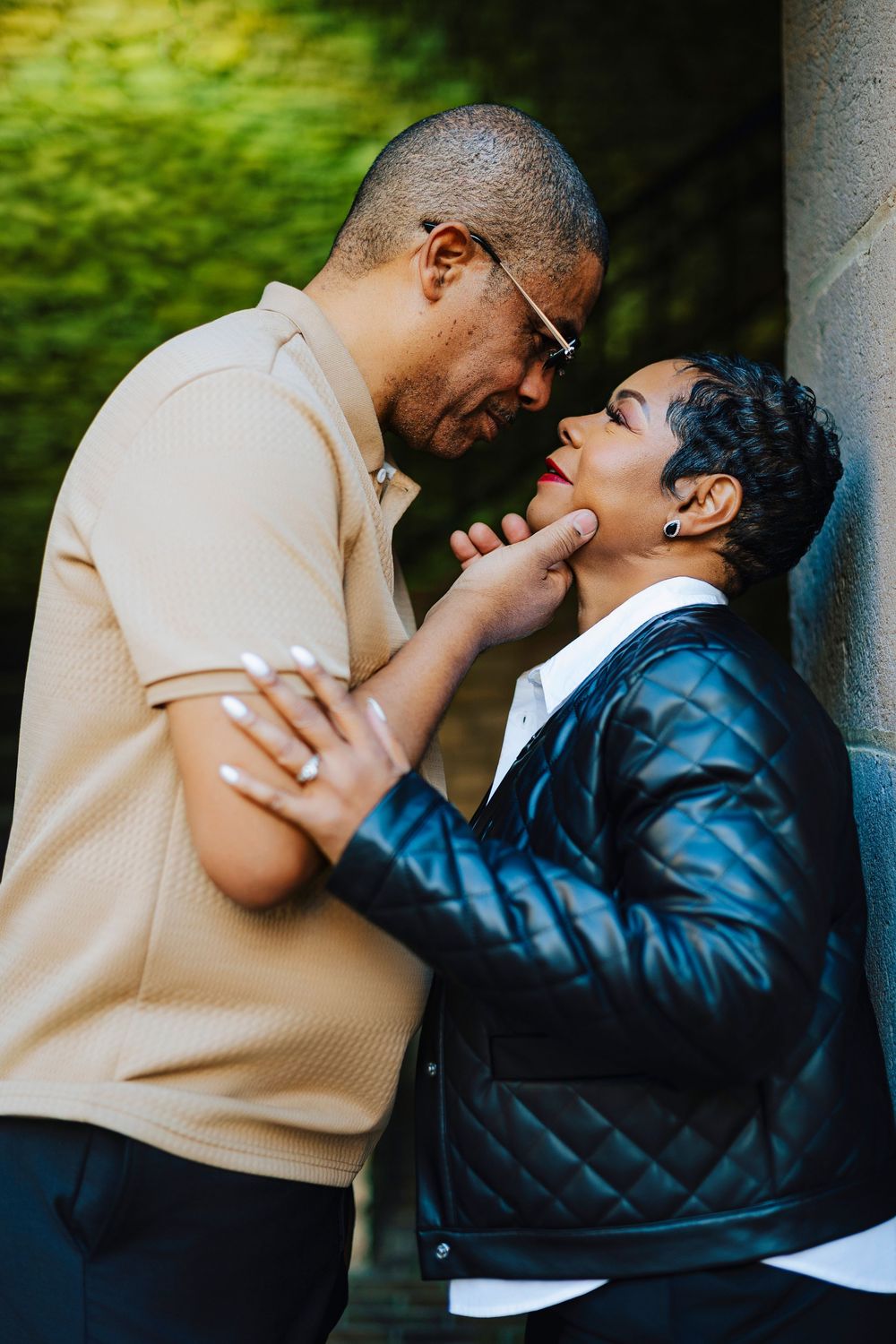 A couple shares a tender moment, foreheads touching, as the man gently cups the woman's face near a stone wall.