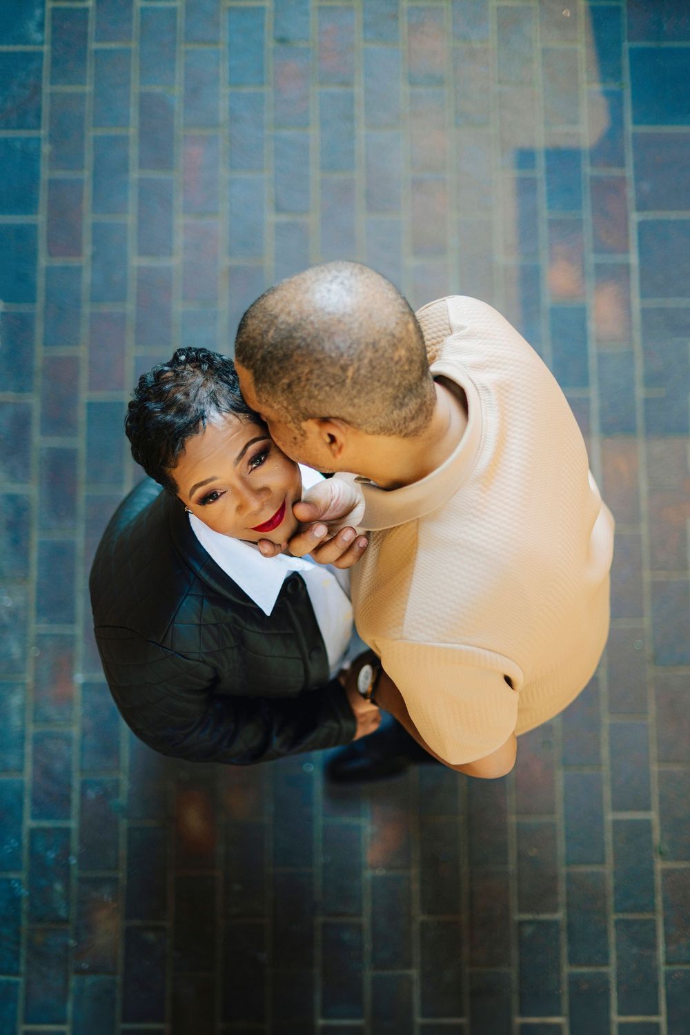 Aerial view of a couple embracing on a brick floor, the man kissing the woman's cheek as she smiles warmly.