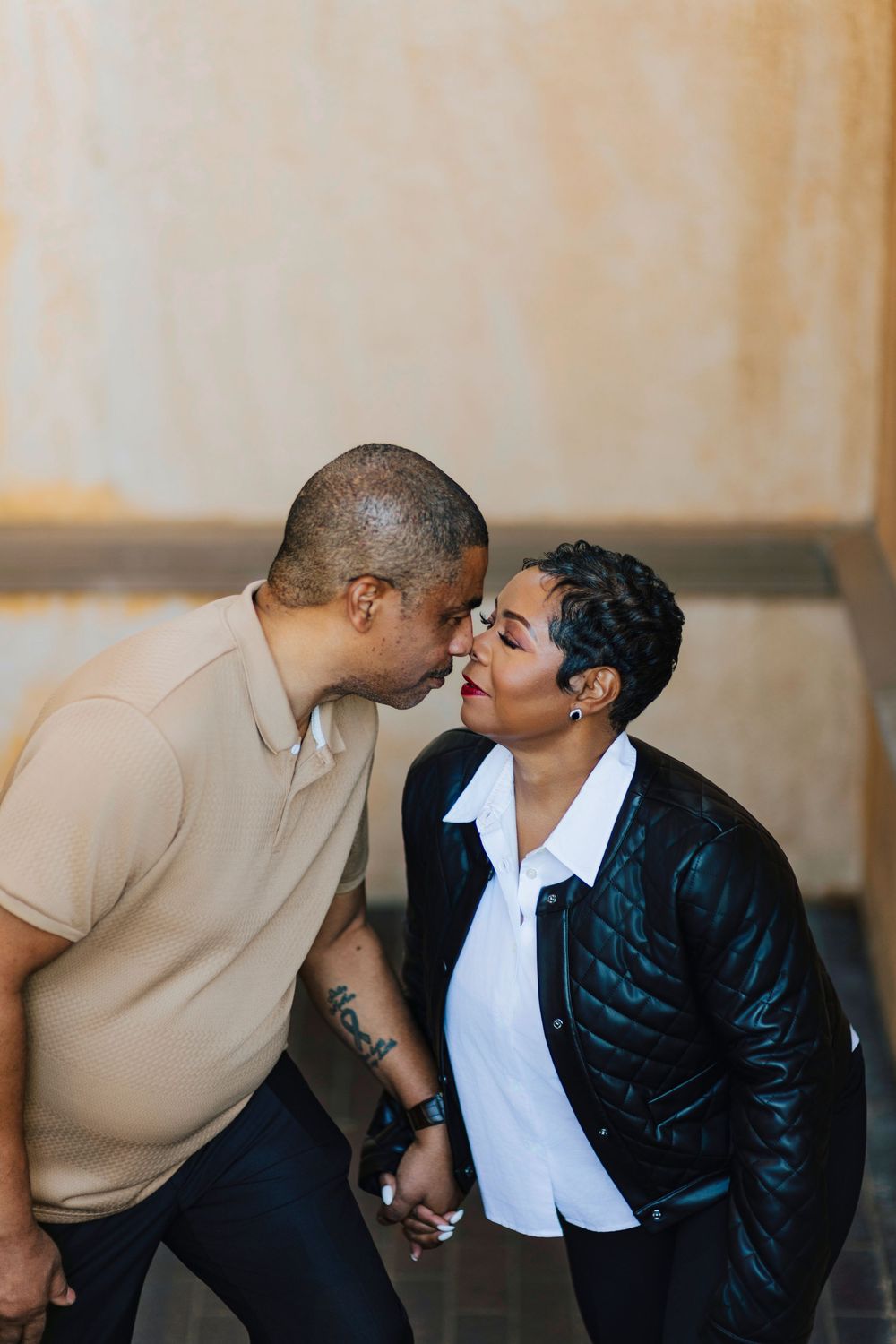 A couple sharing a tender nose-to-nose moment, holding hands, dressed in casual attire against a warm beige wall.