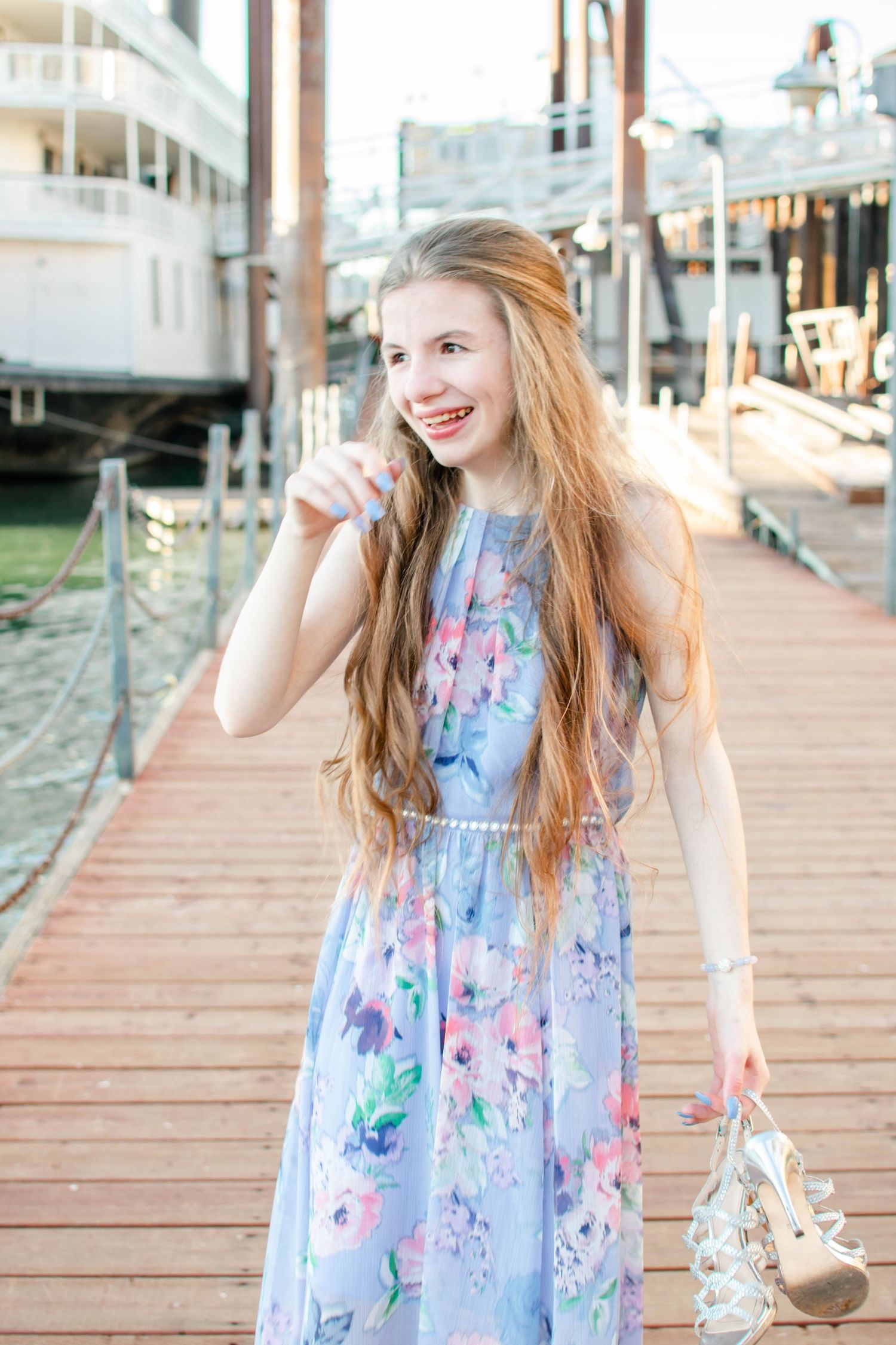 Young woman in floral blue maxi dress holding heels, smiling on a sunny marina boardwalk.