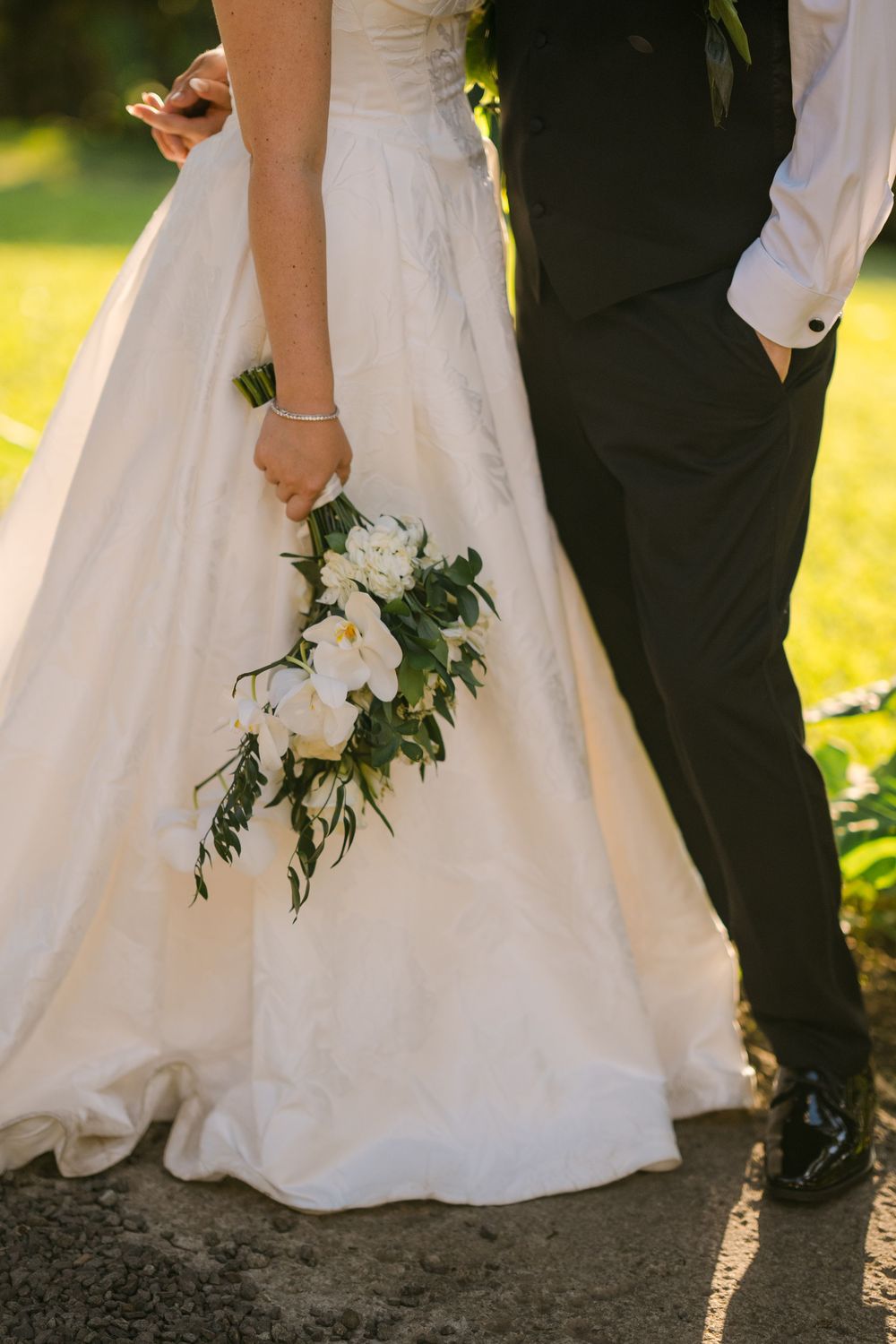 Bride in white gown holding white floral bouquet beside groom in dark suit on sunny wedding day.
