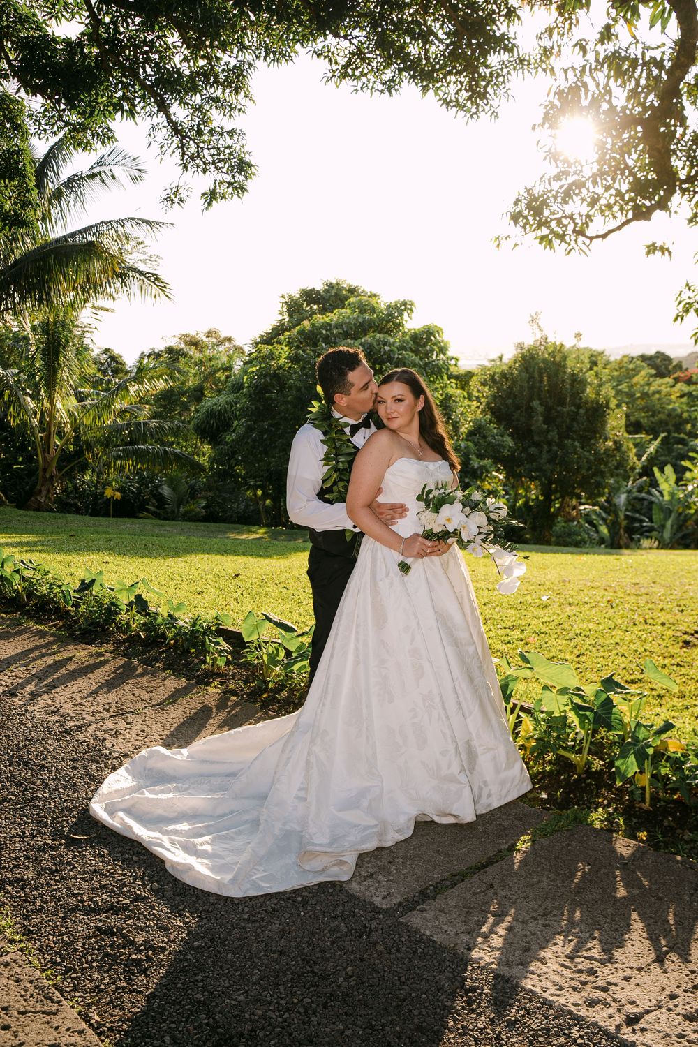 Bride and groom embrace in lush tropical garden at golden hour, bride holding white bouquet in elegant strapless ball gown.