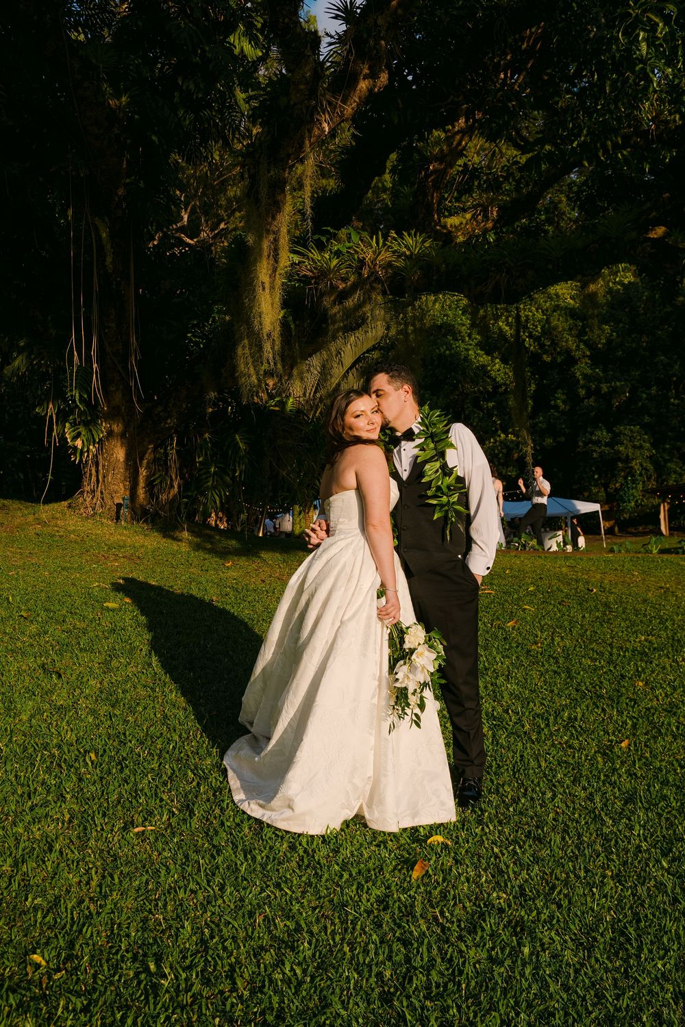 Bride and groom share a romantic kiss on lush green lawn with tropical trees at golden hour wedding.