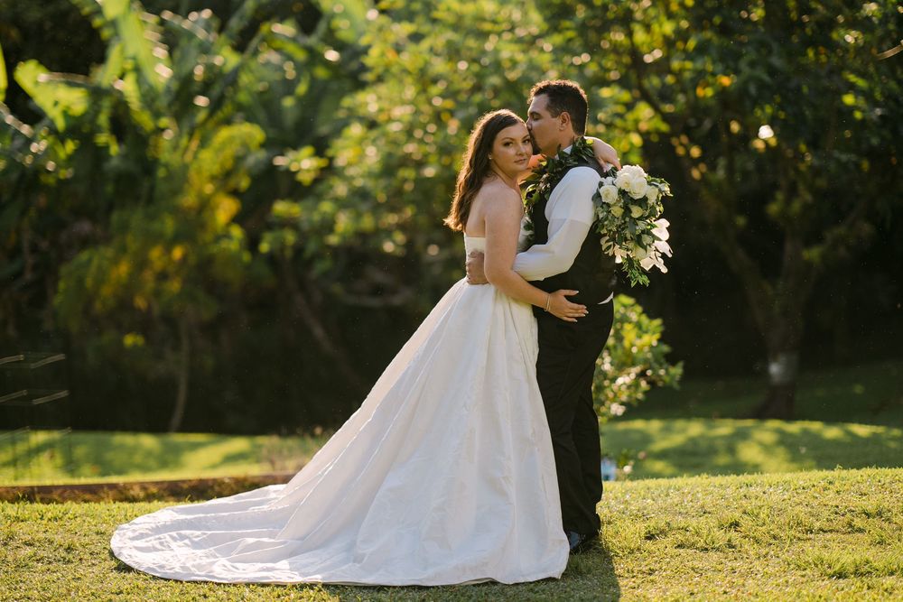 Bride and groom embrace in a sunlit garden, she wears a flowing white ballgown holding a lush bouquet.