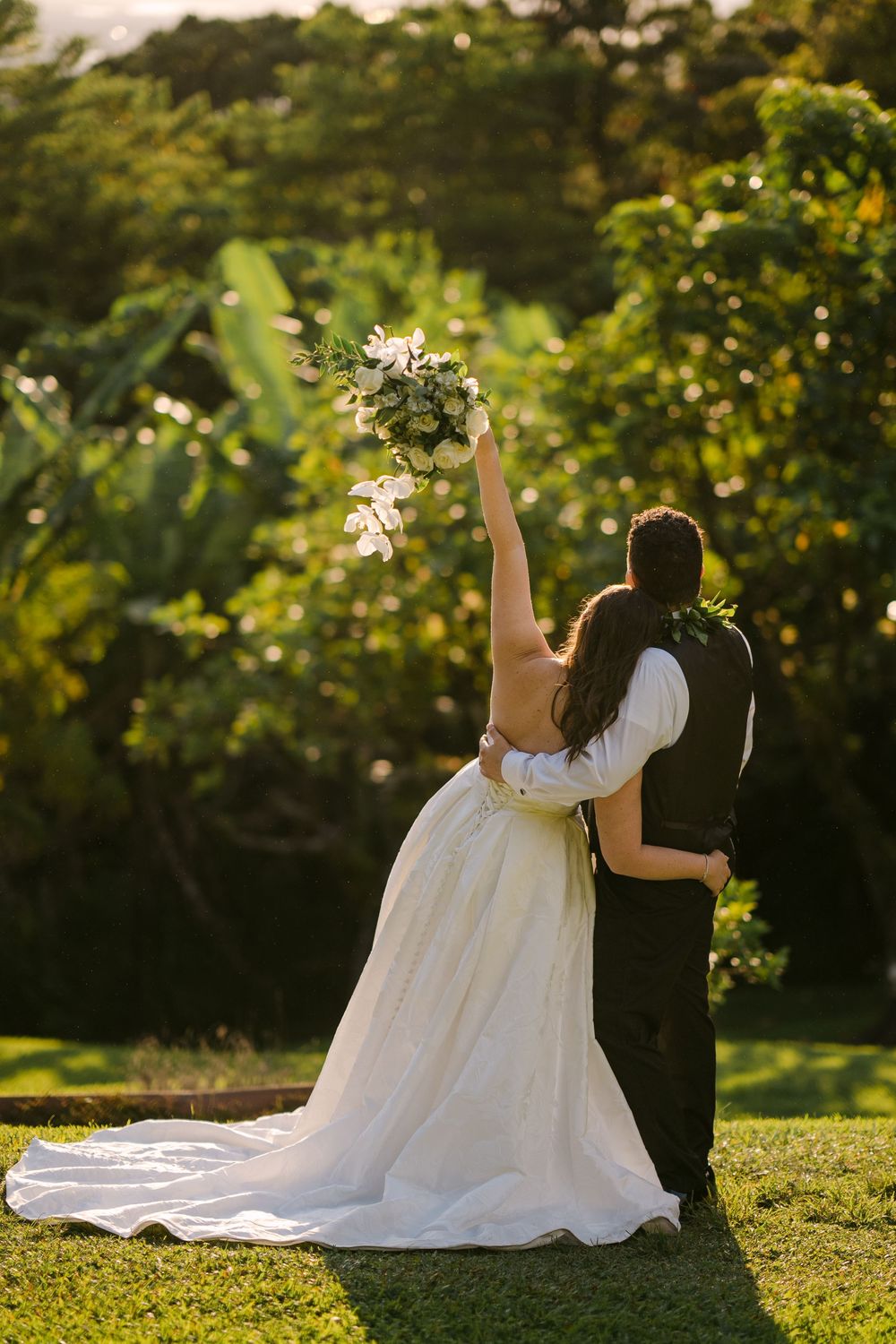 Bride raises bouquet while embracing groom, both facing lush tropical garden backdrop in golden sunset light.