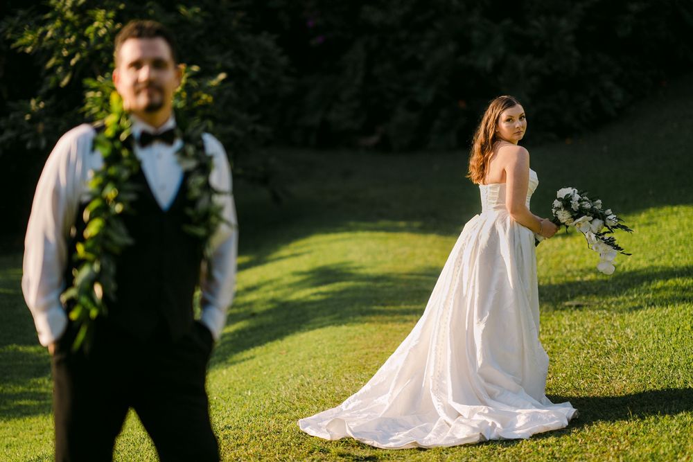 Bride in white gown holds bouquet while groom in black vest with floral lei stands in lush green garden.