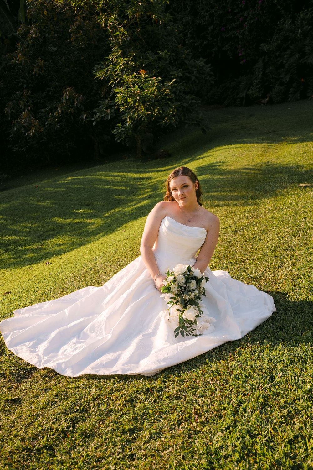 Bride in strapless white ball gown sits on green grass holding white floral bouquet in sunny garden.
