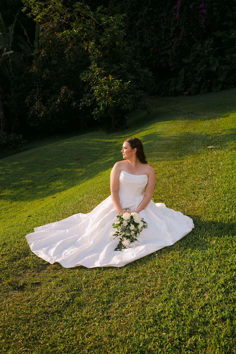 Bride in white strapless gown sitting on green grass holding bouquet of white roses, surrounded by lush garden trees.