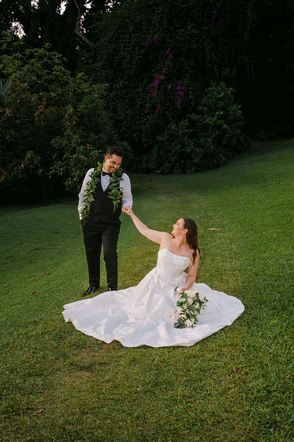 Bride in white gown sits on green grass holding groom's hand in black suit with floral lei, lush garden backdrop.