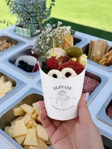 Hand holding an Elevate Charcuterie Bar cup filled with meats, cheeses, olives, raspberries, and floral garnishes.