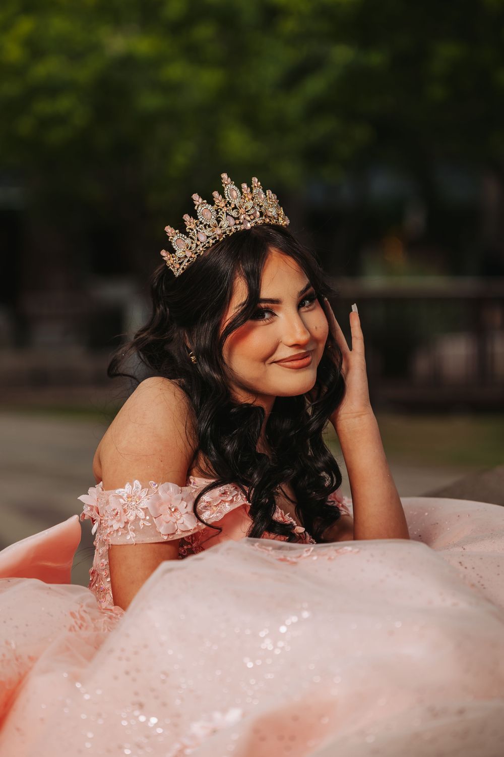 Young woman in pink quinceañera gown and gold tiara poses gracefully outdoors.