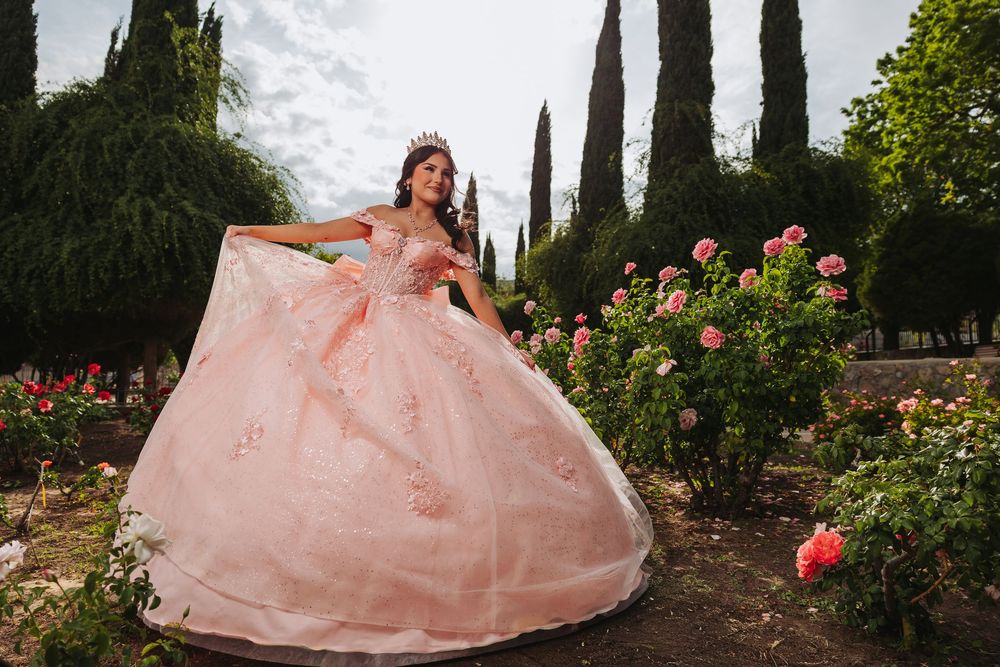 Young woman in a pink ball gown and tiara poses gracefully in a rose garden for her quinceañera.