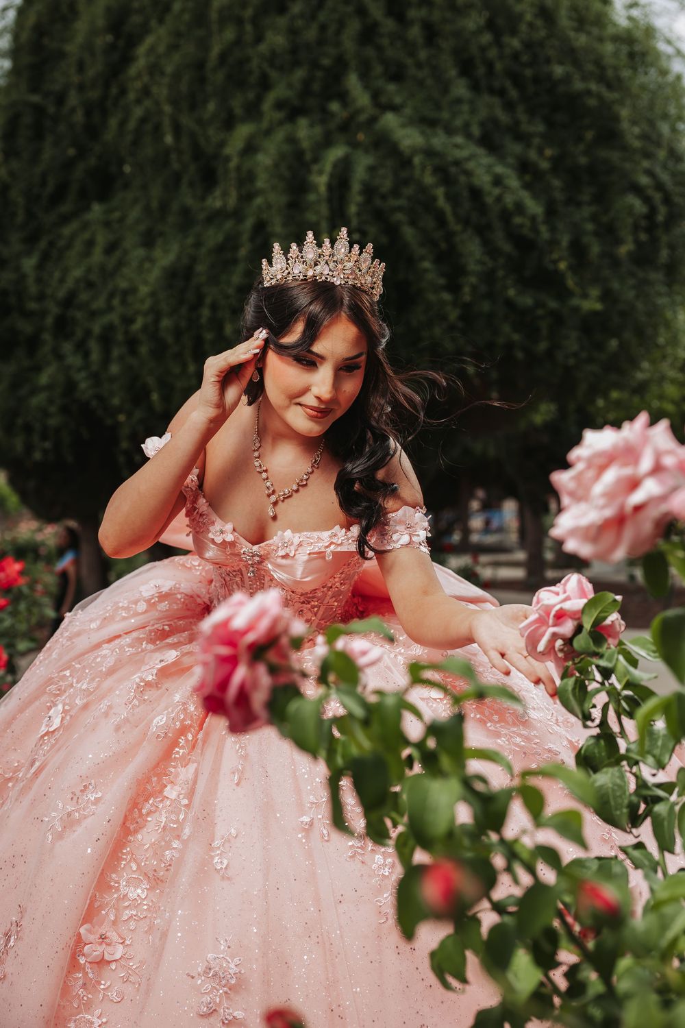 Young woman in pink ball gown and crown poses gracefully among blooming roses in a lush garden setting.
