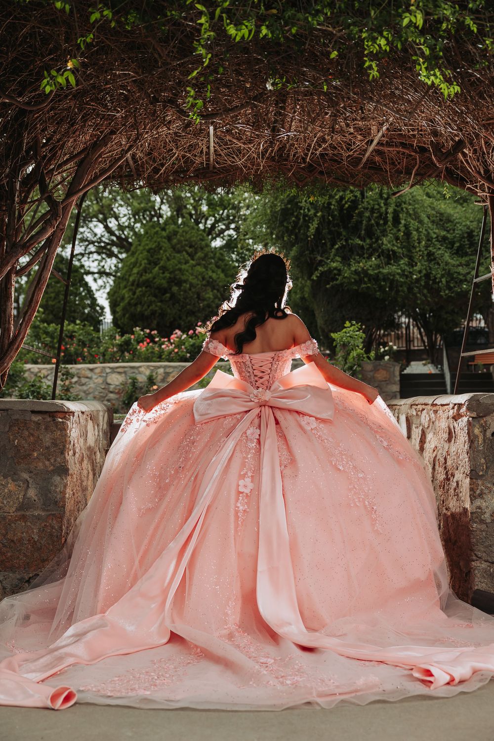 Young woman in a stunning pink ball gown with bow detail and floral lace, posing under a garden arbor.