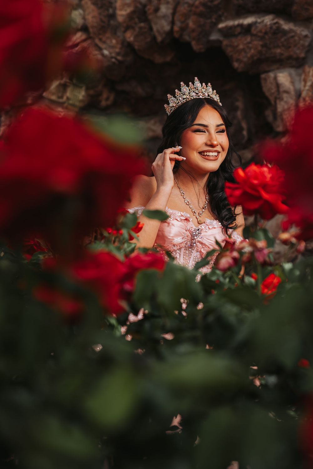 Smiling young woman wearing a crystal tiara and pink gown surrounded by vibrant red roses in a garden setting.