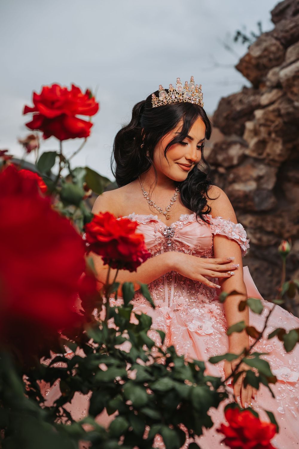Young woman in pink ball gown and gold crown poses among red roses for a quinceañera portrait.