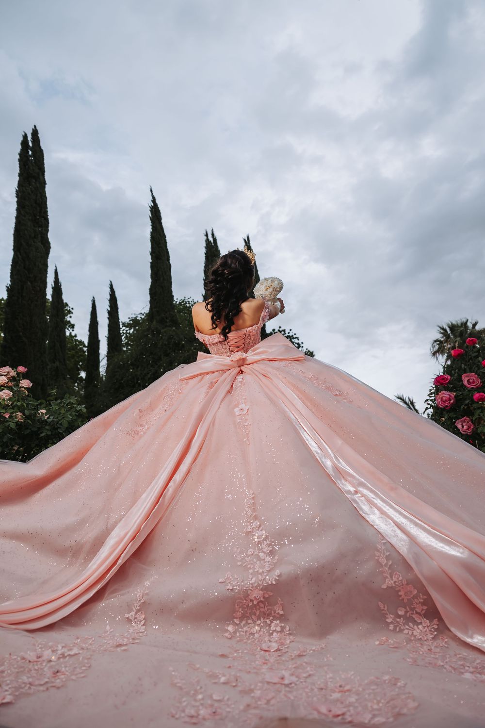 Young woman in pink ball gown quinceañera dress with floral lace, viewed from behind in garden setting.