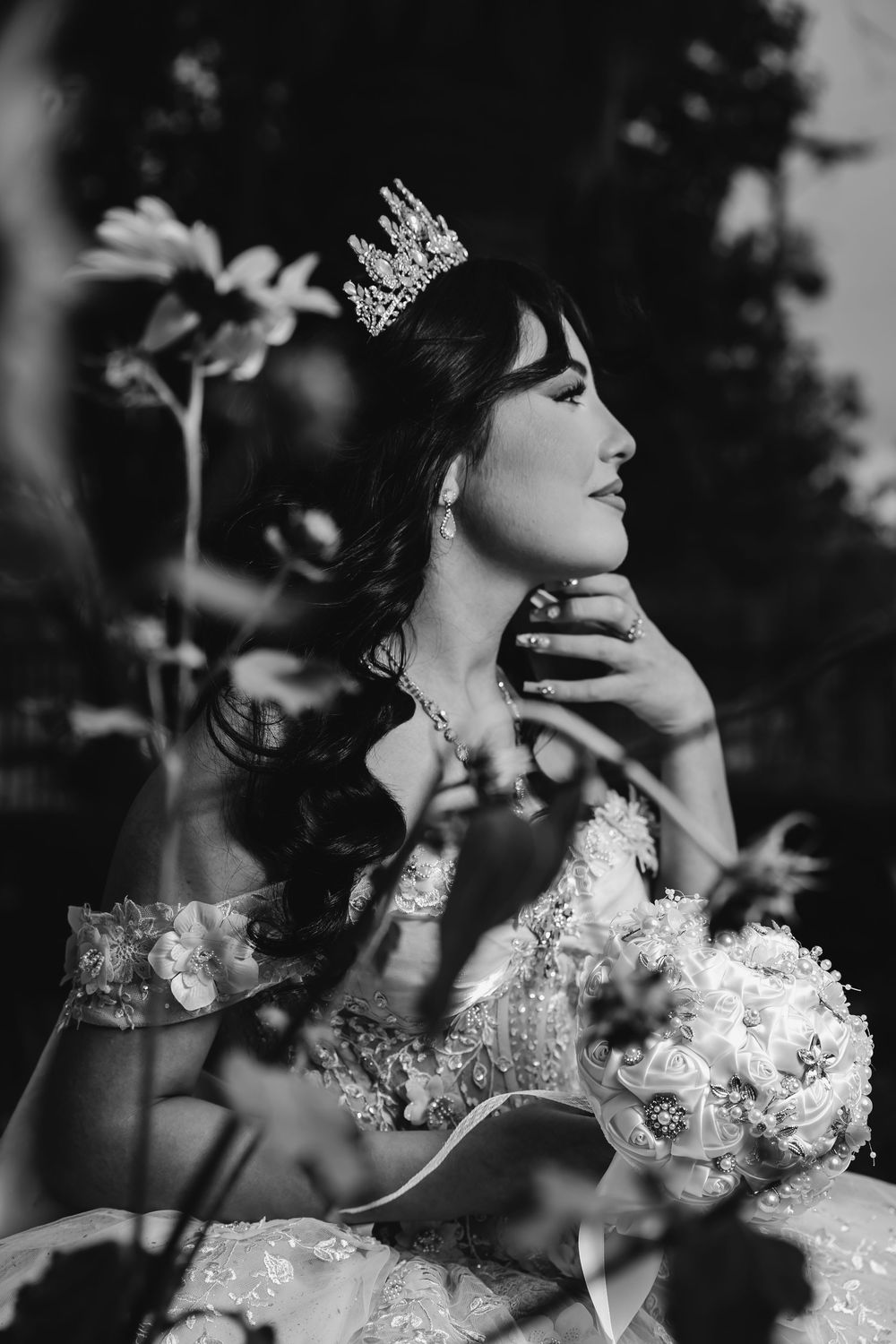 Black and white portrait of elegant bride wearing crown and floral gown, holding bouquet, gazing upward outdoors.