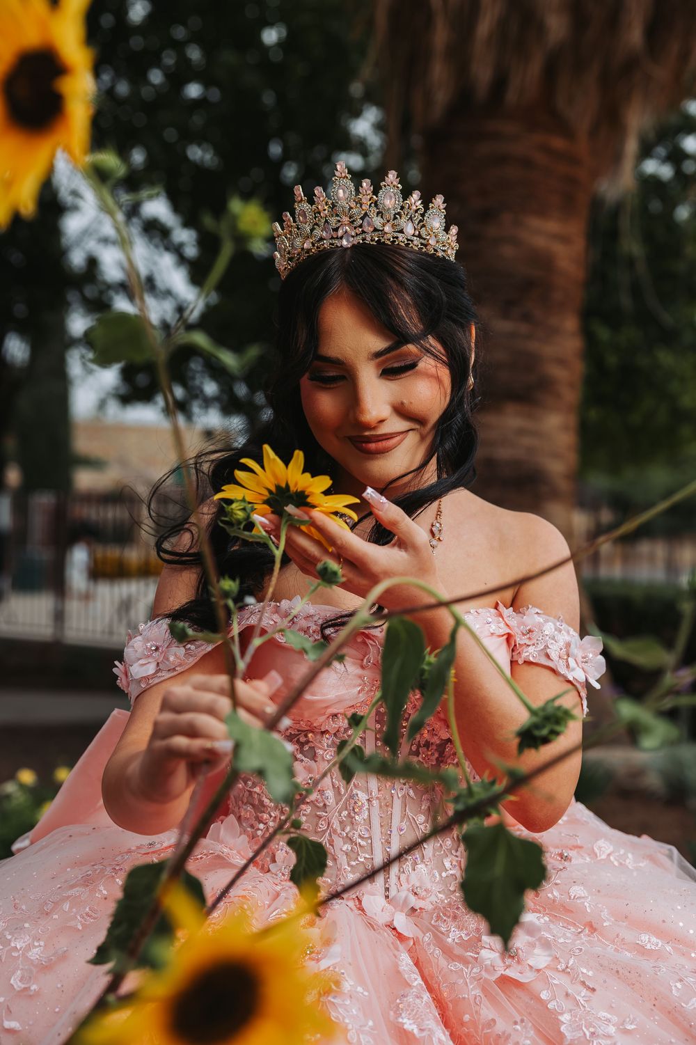 Young woman in pink quinceañera gown and gold tiara crown smiling while touching sunflowers outdoors.