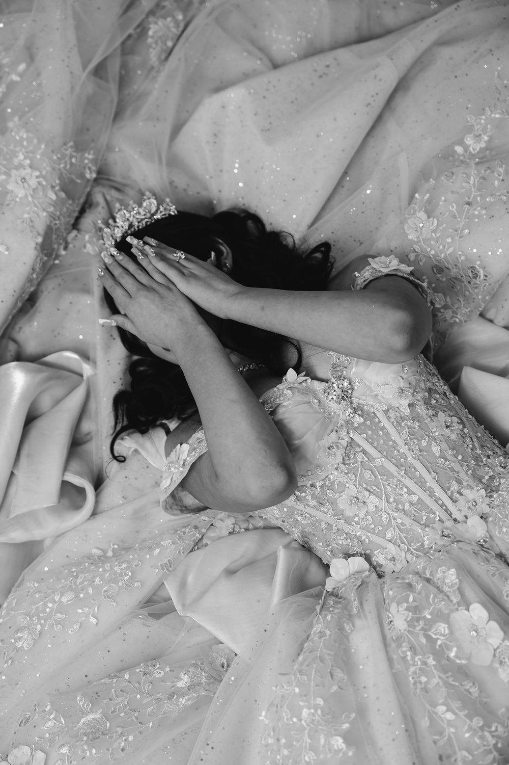 Black and white photo of a bride in an ornate wedding gown lying down, covering her face with her hands.