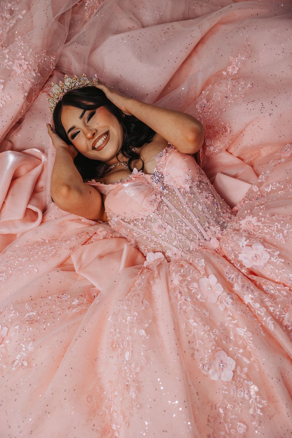 Young woman in pink quinceañera ballgown with crown smiles while lying in her voluminous sparkling dress.