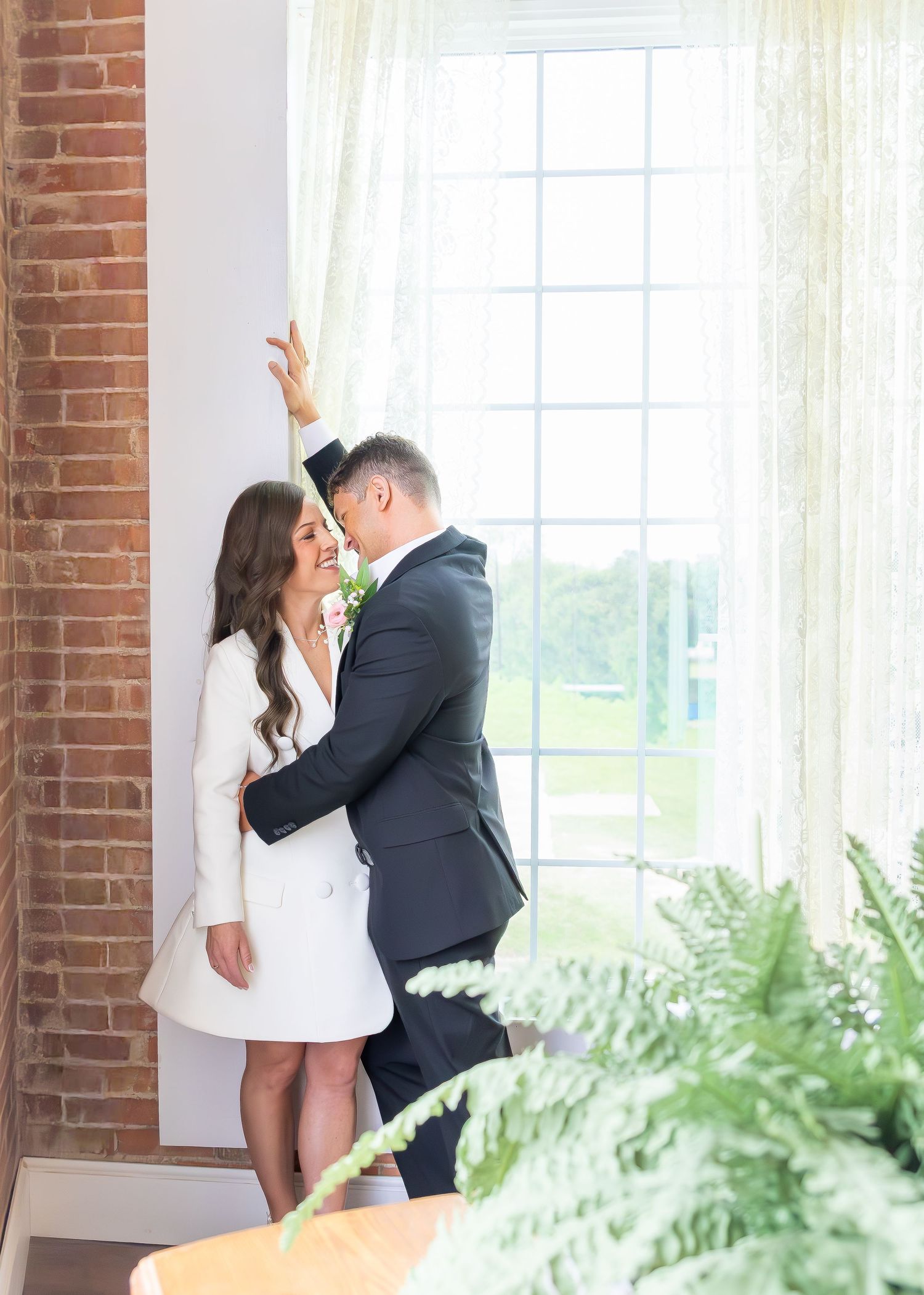 Bride in white mini dress and groom in dark suit sharing romantic moment by sunlit window.