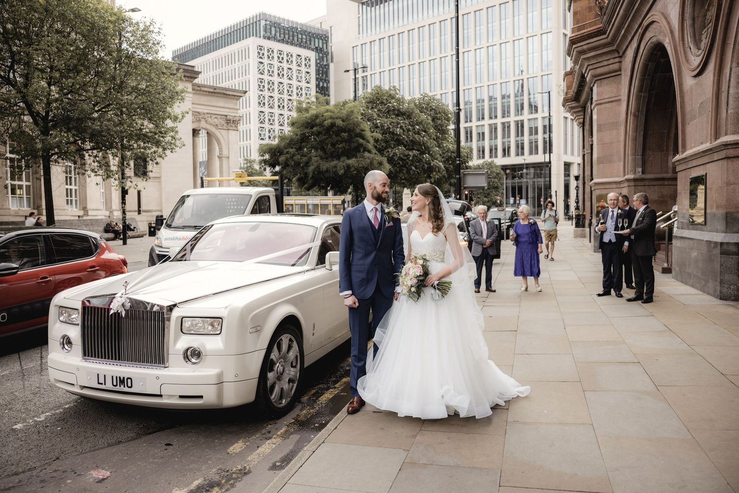 Bride and groom posing beside a white Rolls-Royce Phantom outside a city venue on their wedding day.