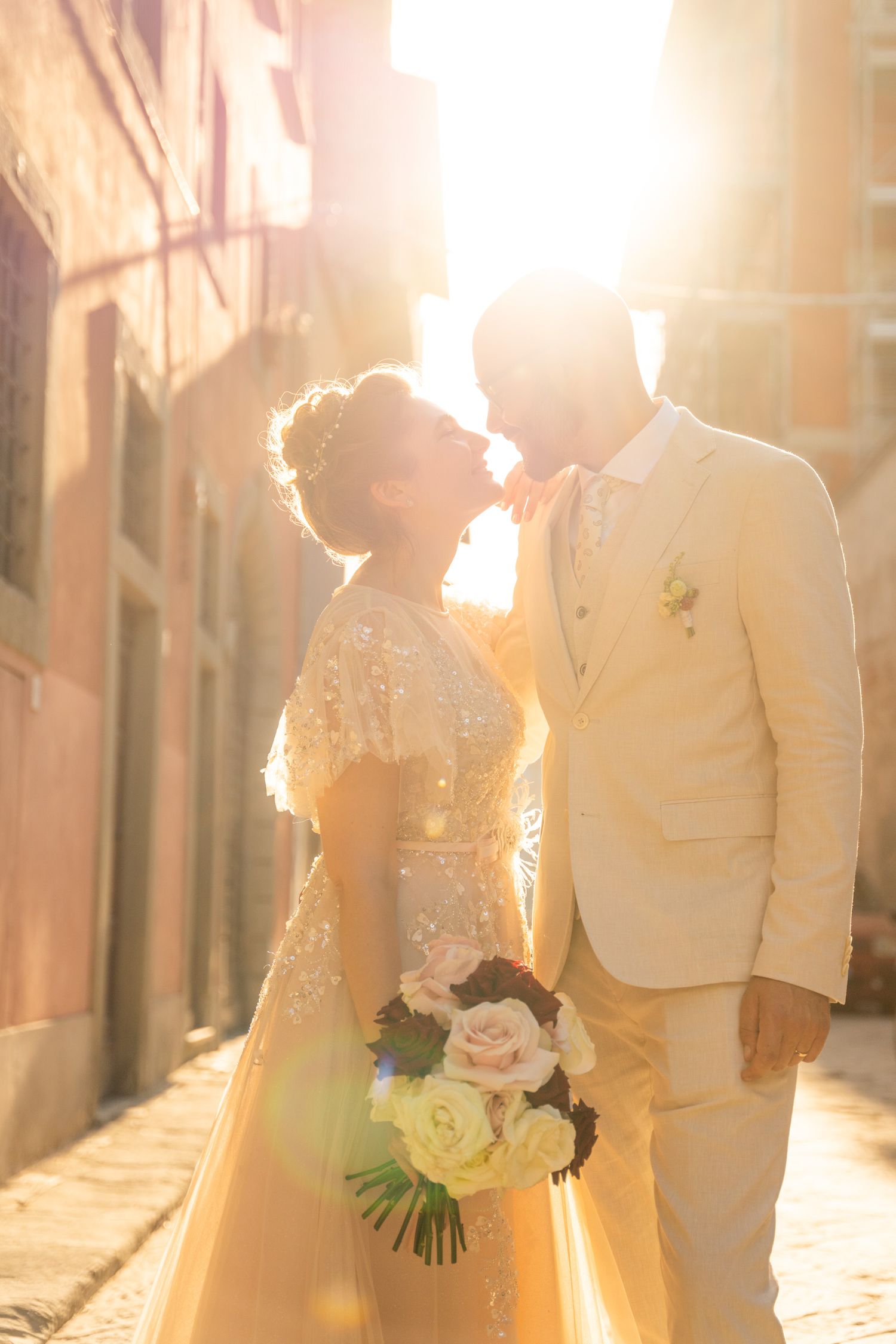 A bride and groom share a romantic kiss in a sunlit cobblestone alley, bride holding a floral bouquet.