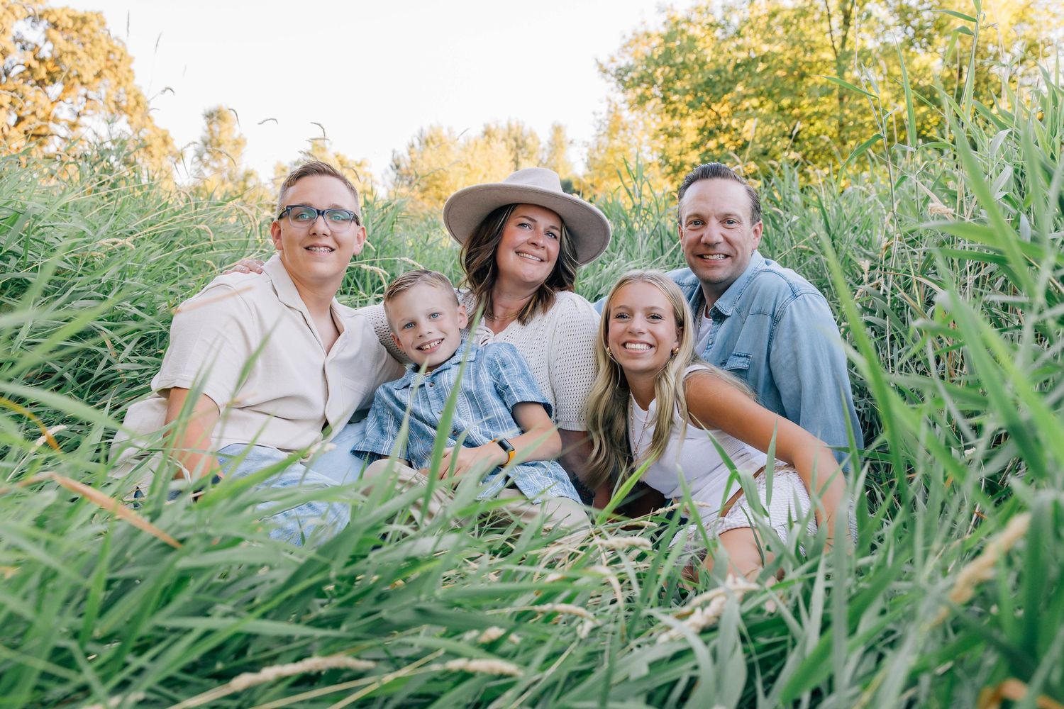 Happy family of five smiling together in lush green tall grass field during golden hour outdoor photo session.