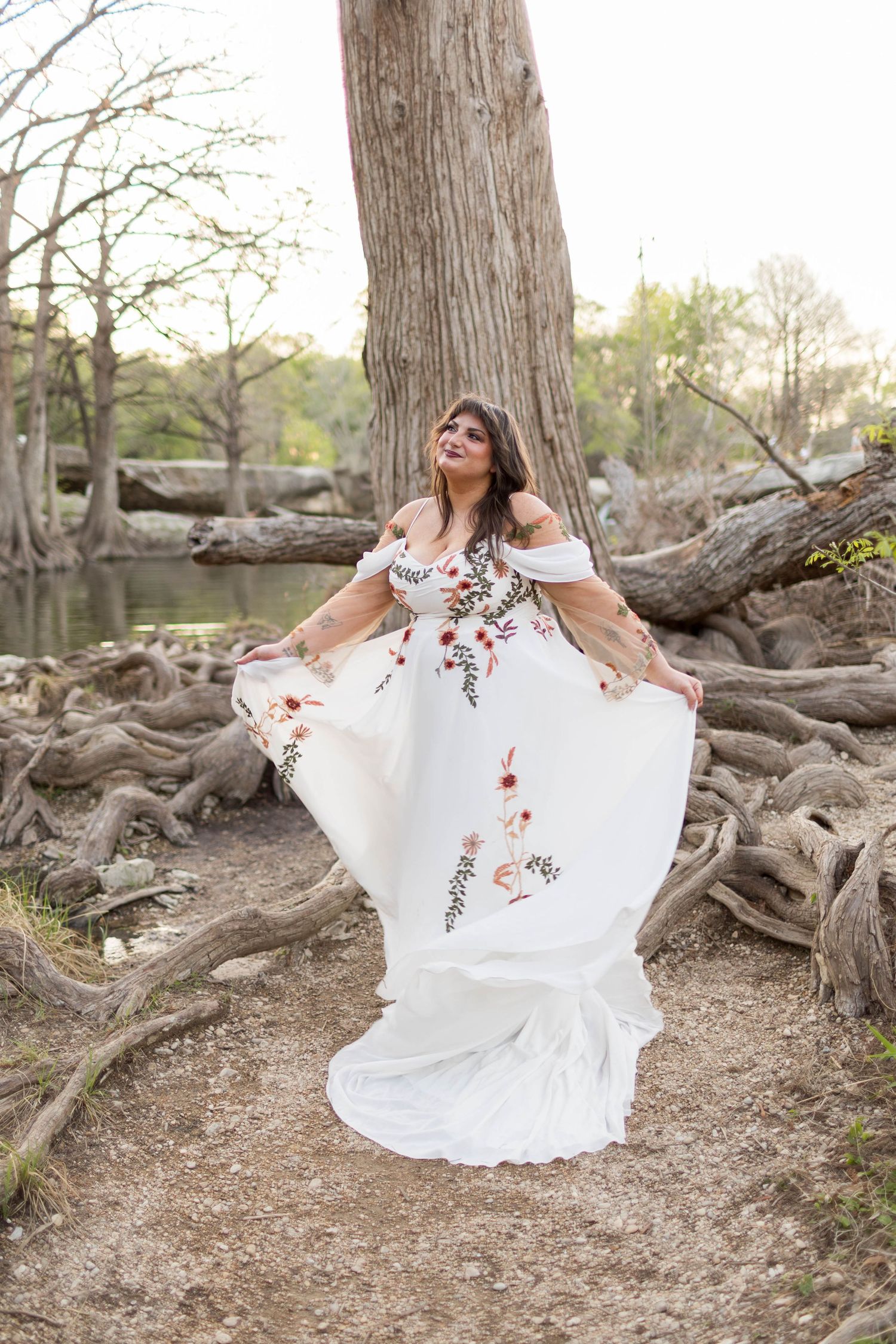 Woman in white floral off-shoulder gown twirling near tree roots by a river in a dreamy outdoor portrait in Austin, Texas.