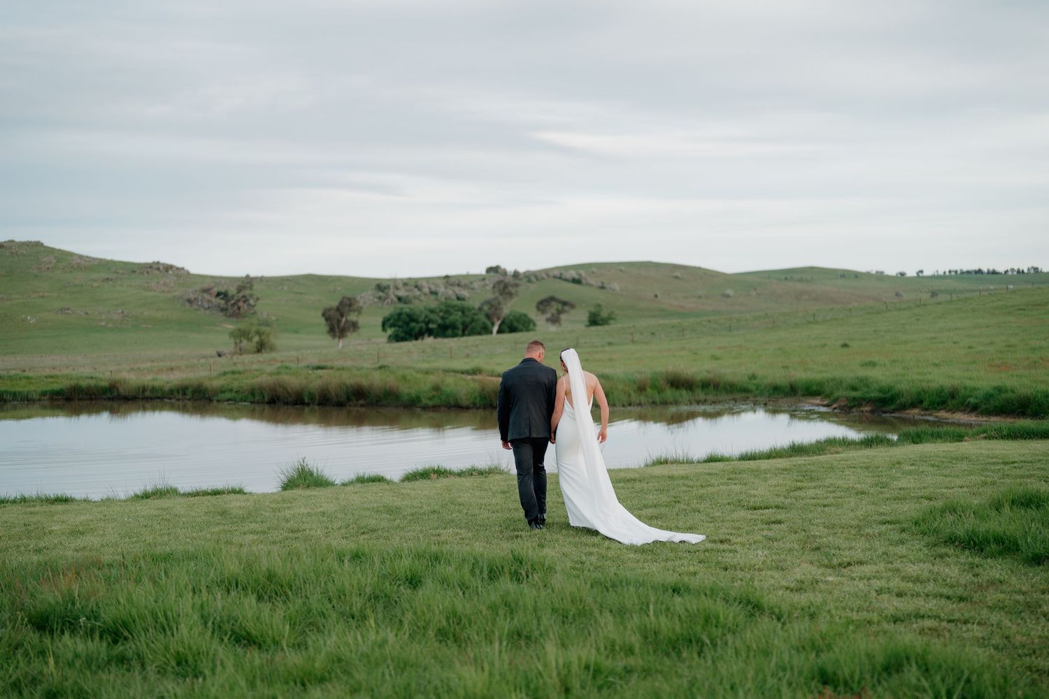Bride and groom walking hand in hand through lush green rolling hills beside a serene pond on their wedding day.