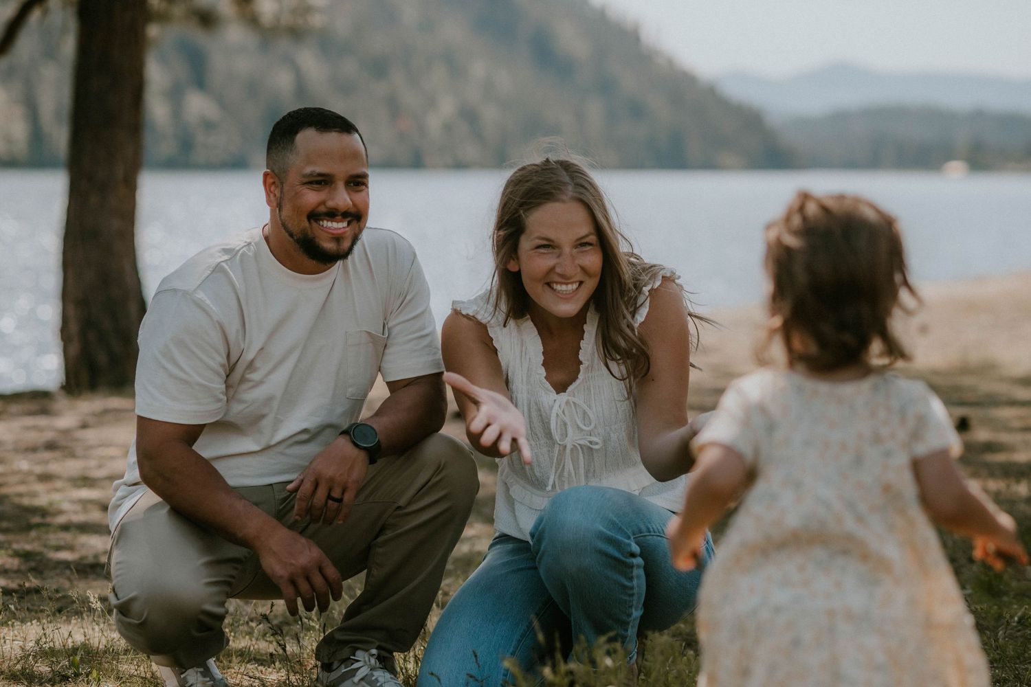 A couple smiles warmly at their toddler daughter outdoors near a lake surrounded by trees.