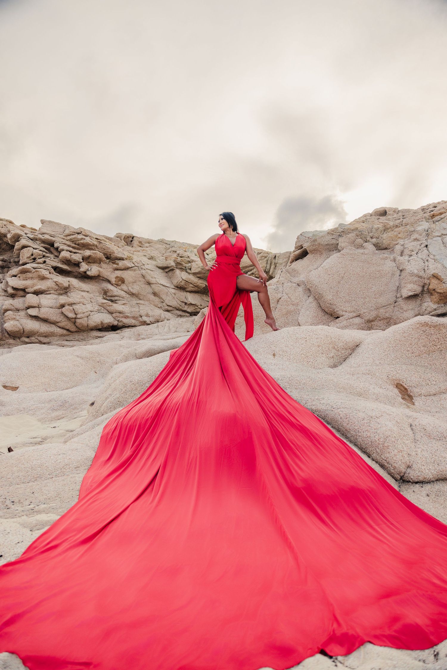 Woman in flowing red gown with dramatic train poses on rocky desert landscape.