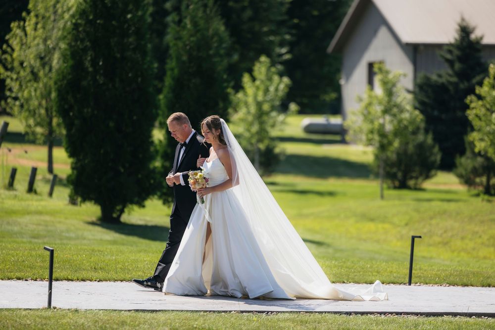 Bride in white gown and veil walks with her father down the aisle near white chapel in Wisconsin on sunny wedding day.