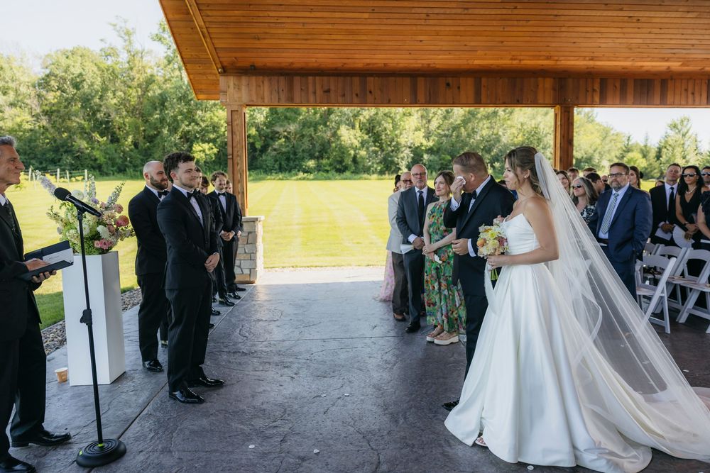Outdoor Wisconsin wedding ceremony under a pavilion with bride in white gown and veil facing groom in black tuxedo.