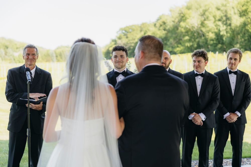 Outdoor Wisconsin vineyard wedding ceremony with bride in white veil facing groom and groomsmen in black tuxedos on a sunny day.